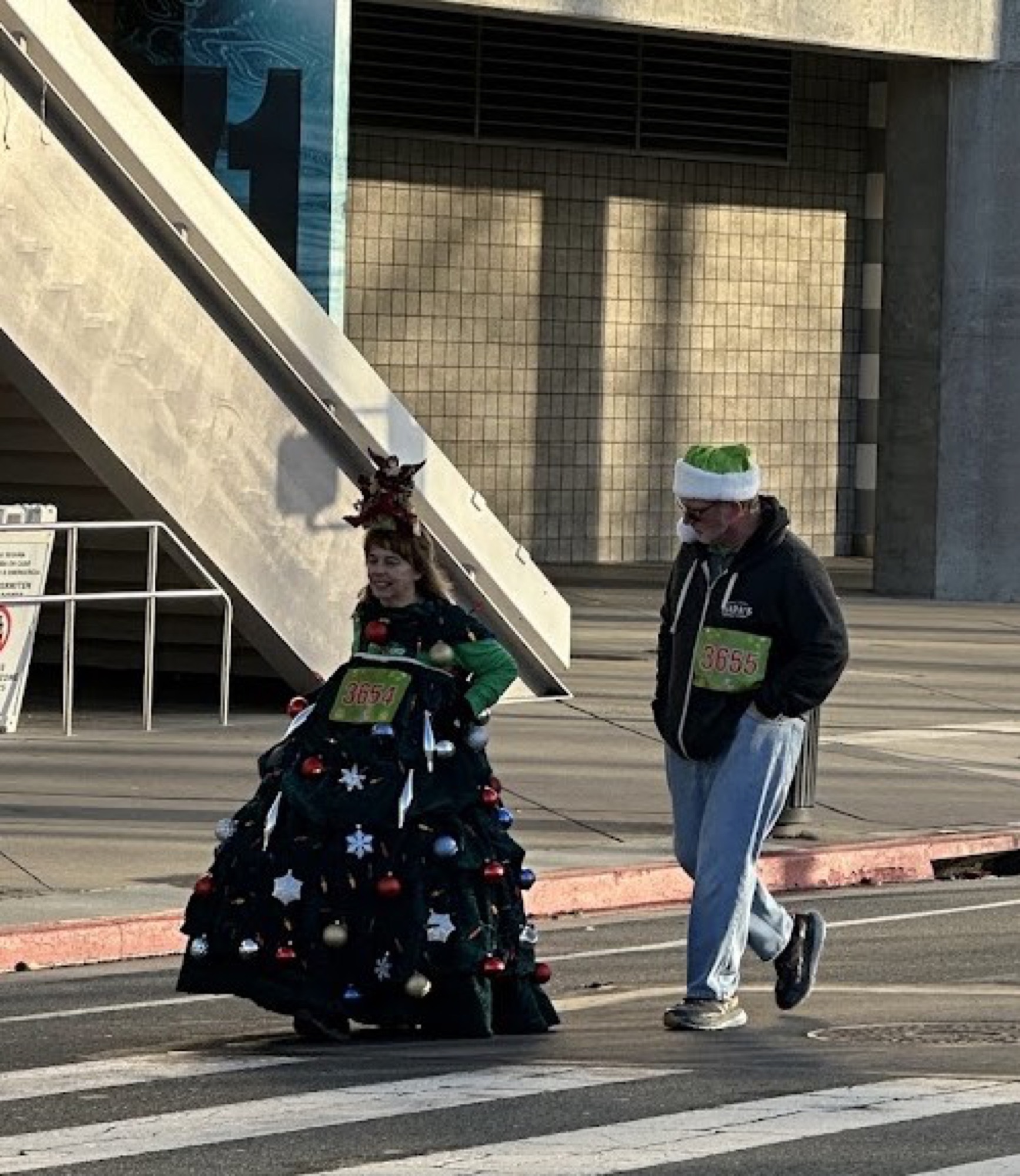 A participant dressed as a decorated Christmas tree walking with a friend in an elf hat